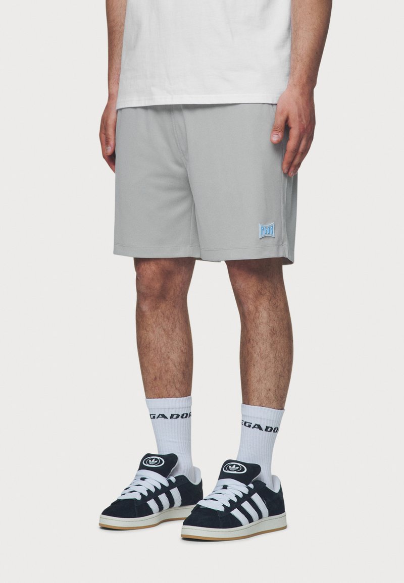 Grå treningshorts med elastisk midje og en blå logo-patch. kombinert med hvite sokker og svarte semskede sneakers med striper.