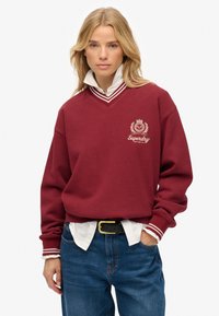 Sudadera burdeos con escote en V y acentos a rayas en blanco y negro. Presenta un logo bordado en el pecho. Lleva puesta sobre una camisa con cuello.