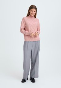 Un pull en tricot rose clair avec un col montant, présentant un motif en losanges et des poignets côtelés, associé à un pantalon large gris et des chaussures noires.