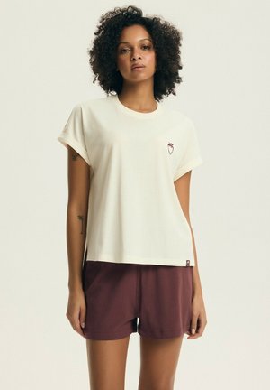 Kobieta o kręconych włosach ubrana w białą koszulkę z krótkim rękawem z małym wzorem truskawki i bordowe szorty, stoi na jednolitym tle.