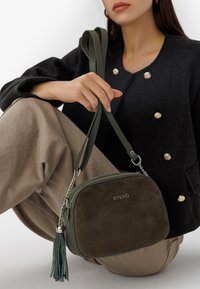 Piccola borsa a tracolla verde oliva in morbida suede, con hardware argentato, tracolla a catena e un accento con nappina. Logo del brand impressa sul davanti.