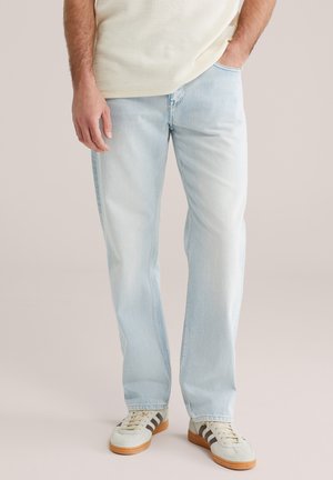 Lichtblauwe spijkerbroek met rechte pijpen, een vervaagde afwerking en subtiele strepen. Gecombineerd met witte sneakers met bruine strepen.