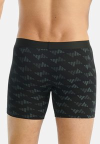 Muž nosící černé boxerky s tmavě šedým geometrickým trojúhelníkovým vzorem, zobrazený od dolní části zad po střed stehna.