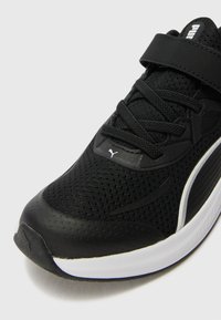 Chaussure de sport noire avec tige en mesh, embout renforcé, sangle ajustable et semelle blanche avec des accents texturés.