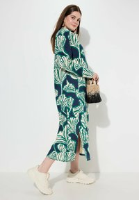 Maxiklänning med blommönster i marinblått och grönt, med långa ärmar och sprund på sidorna. Matchad med en handväska i svart och beige samt vita sneakers.