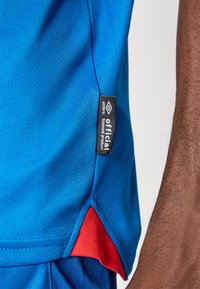 Blaues Sportshirt mit Mesh-Struktur, seitlichem Schlitz mit rotem Akzent und einem schwarzen Etikett mit der Aufschrift "offizielles lizenziertes Produkt" von Umbro.