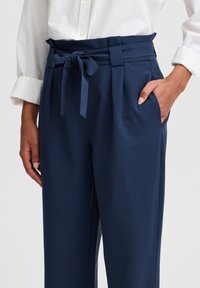 Pantalons taille haute bleu marine avec une ceinture nouée, dotés d'une coupe large et de poches latérales. Tissu lisse avec une finition soignée.