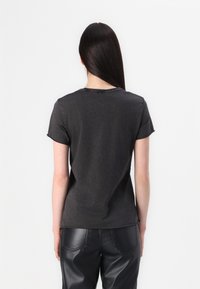 Kvinde med langt mørkt hår iført en ensfarvet mørkegrå T-shirt og sorte læderbukser, vendt væk fra kameraet mod en hvid baggrund.