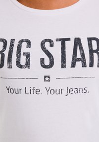 Bílá košile s opotřebovaným černým nápisem "BIG STAR" a menším textem pod ním, který uvádí "Your Life, Your Jeans."