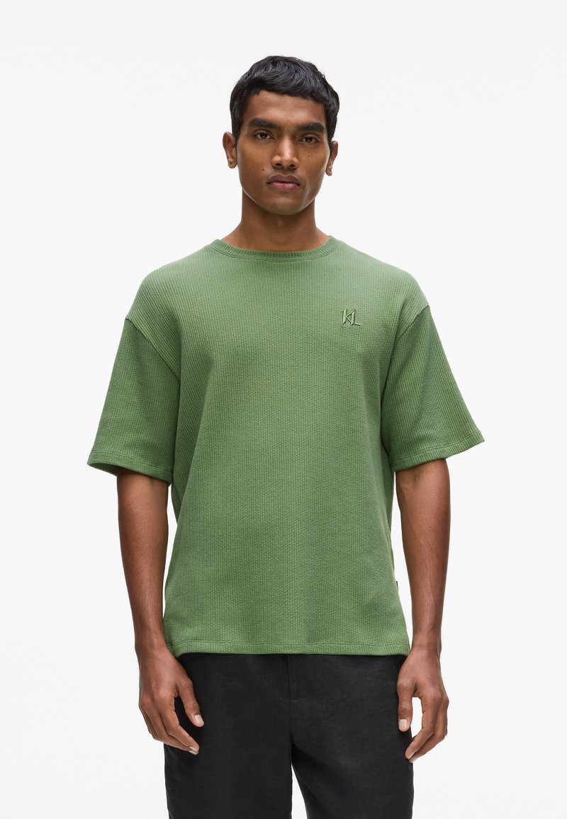 Chemise verte en maille gaufrée à manches courtes avec un col rond et un logo brodé sur la poitrine. Portée avec un pantalon foncé et ample.