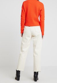 Lyhennetty, teksturoitu oranssi neulepaita pitkillä hihoilla yhdistettynä valkoisiin leveisiin farkkuihin, joiden helmassa on purjehtiminen, ja mustiin korkeakorkoisiin saappaisiin.