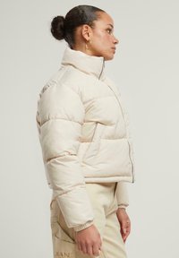 Lys beige pufferjakke med høy krage, glidelås foran og cropped design. Har sømte seksjoner og elastiske mansjetter. Matt finish.