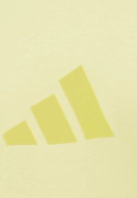 Světle žlutá látka s hladkou strukturou obsahuje ikonické logo se třemi pruhy ve slabě tmavší žluté, umístěné diagonálně.