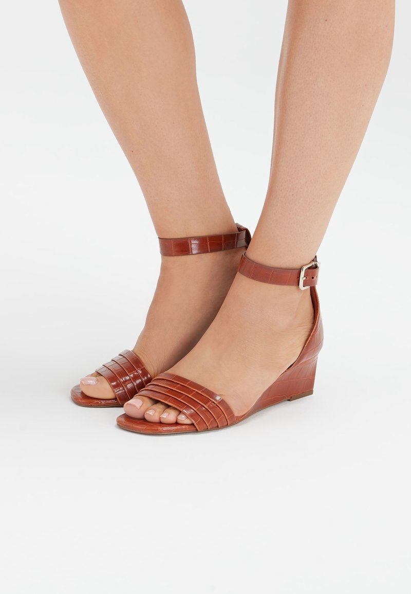 Pieds chaussés de sandales compensées en cuir marron avec brides à la cheville et orteils découverts sur fond blanc uni.
