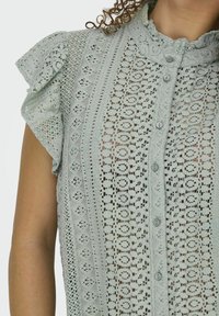 Lichtgroene kanten blouse met korte ruches bovenstukken, knoopsluiting en ingewikkelde geweven patronen met cirkels en lineaire ontwerpen.