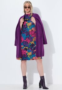 Fioletowy sweter z dzianiny noszony na kolorowej sukience w kwiaty w odcieniach niebieskiego, różowego i zielonego, zestawiony z czarnymi botkami za kostkę.