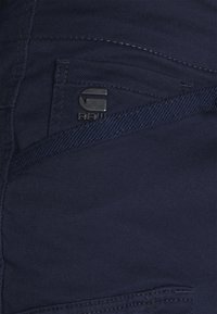 Dunkelblaue Denim-Stoff mit strukturiertem Finish, mit einem sichtbaren Logo-Tag und kontrastierenden Nähten im Taschenbereich.