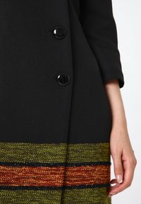Svart kappa med två blanka knappar, med en texturerad kant i gröna, orange och svarta ränder. Slät tyg, skräddarsydd design.