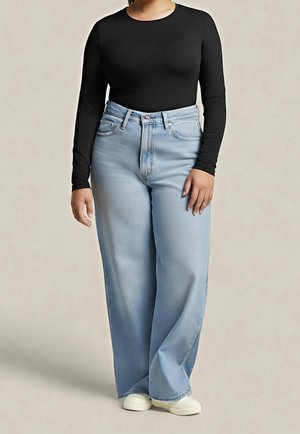 Vrouw staand, gekleed in een nauwsluitende zwarte top met lange mouwen, lichtblauwe wijd uitlopende spijkerbroek en witte sneakers, tegen een effen achtergrond.