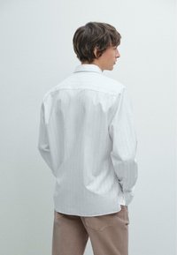 Weißes Langarmhemd mit schwachen vertikalen Streifen, Button-Down-Kragen und Manschetten in Fassform, kombiniert mit hellbraunen Hosen. Glatte Textur.