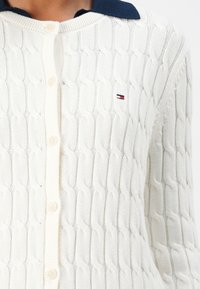 Hvit kabelstrikket cardigan med marineblå krage, knappestolpe og liten brodert logo på venstre bryst. Myk tekstur, lange ermer.