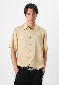 Camisa de manga corta, color amarillo claro, con una textura sutil, botones frontales y un logotipo bordado. Combinada con un cinturón negro que presenta una hebilla de plata.