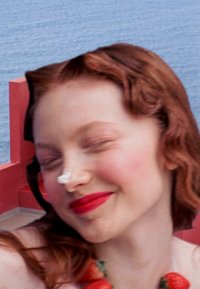 Junge Person mit roten Locken, trägt ein weißes Oberteil und eine Erdbeeren-Halskette, lächelt mit Sahne auf der Nase vor einem blauen Ozeanhintergrund.