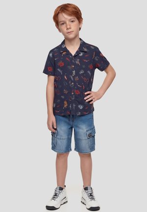 Niño pelirrojo de pie con la mano en la cadera, vistiendo una camisa azul marino con estampado floral de manga corta, pantalones cortos tipo cargo de mezclilla y zapatillas blancas sobre un fondo gris.