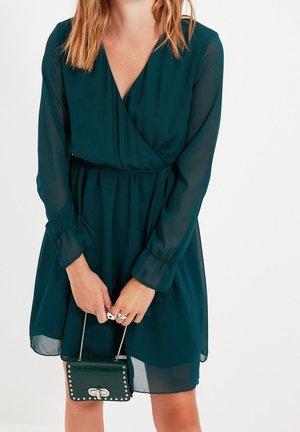 Frau in dunkelgrünem Wickelkleid mit langen Ärmeln, die eine kleine dunkelgrüne Handtasche mit silbernen Nieten und Verschluss hält.