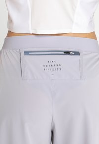 Pantalones cortos de running gris claro con cintura baja, que cuentan con un bolsillo trasero con cremallera y el texto "División de Running de Nike" en la cintura.