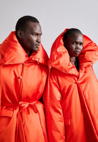 Δύο άτομα φορούν υπερμεγέθη, φωτεινά πορτοκαλί σατέν μπουφάν με μεγάλα καπέλα και δεμένους ζώνες. Τα μπουφάν διαθέτουν κουμπιά κλεισίματος και πλευρικές τσέπες.