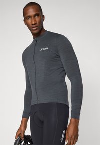 Maillot de cyclisme à manches longues gris foncé avec une fermeture éclair à l'avant et le logo "LE COL". Porté avec un short de cyclisme rembourré noir. Texture de tissu lisse.