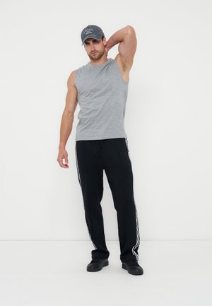 Άνδρας με γκρι αμάνικο πουκάμισο, μαύρο παντελόνι φόρμας με λευκές ρίγες, μαύρα παπούτσια και μπλε καπέλο, στέκεται μπροστά σε λευκό φόντο, με το ένα χέρι πίσω από το κεφάλι.