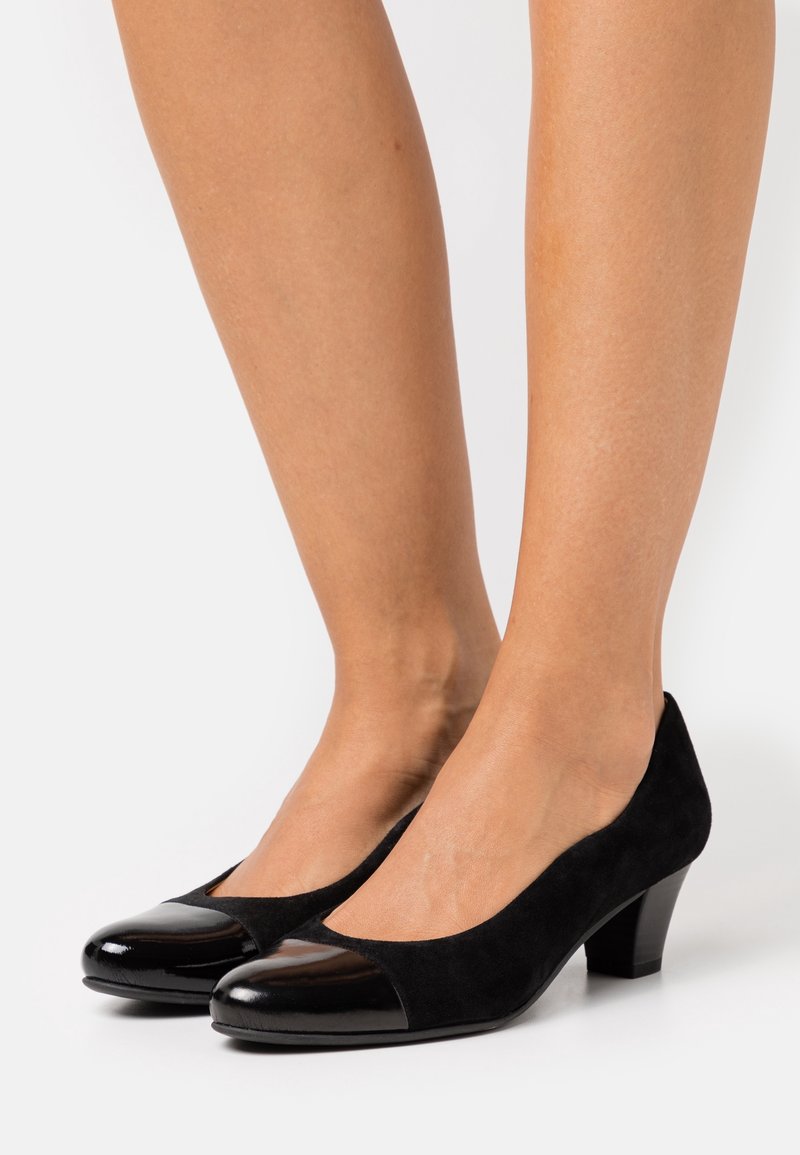 Femme portant des chaussures basses en daim noir avec des bouts ronds noirs brillants, debout sur une surface blanche.