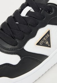 Adidași alb-negru, cu piele texturată, șireturi negre și un logo triunghiular aurit cu "GUESS U.S.A." pe lateral.