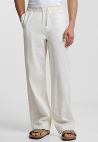 Witte linnen broeken met een elastische tailleband, zijzakken en een relaxte pasvorm. De stof heeft een subtiele textuur, gecombineerd met bruine sandalen.