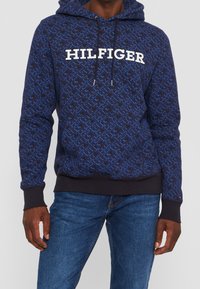Námořnická modrá mikina s kapucí, s potištěným designem, na hrudi velké bílé logo "HILFIGER", kapuce na stažení a žebrované manžety.