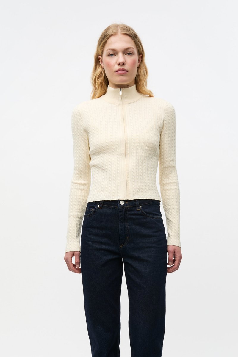 Kremfarget ribbestrikket genser med glidelås, høy krage, tettsittende design og lange ermer, kombinert med mørkeblå høytlivsjeans.