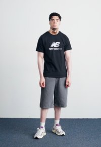 T-shirt nera New Balance con logo, pantaloni corti in denim grigi, calzini bianchi alla caviglia e sneakers grigie e viola. In piedi contro uno sfondo semplice.