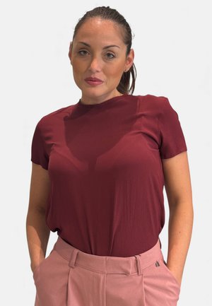 Donna con capelli raccolti che indossa una maglietta bordeaux a maniche corte e pantaloni rosa a vita alta, in piedi con le mani nelle tasche.