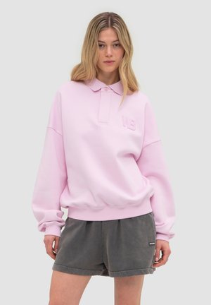 Vrouw draagt een lichtroze polo-sweatshirt met lange mouwen en een "WB"-logo, en losse donkergrijze shorts tegen een effen lichte achtergrond.