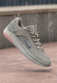 Bež Nike tenisica s mrežastim gornjim dijelom, debelim bijelim potplatom i vezicama, lebdi iznad ružičaste betonske površine na otvorenom.