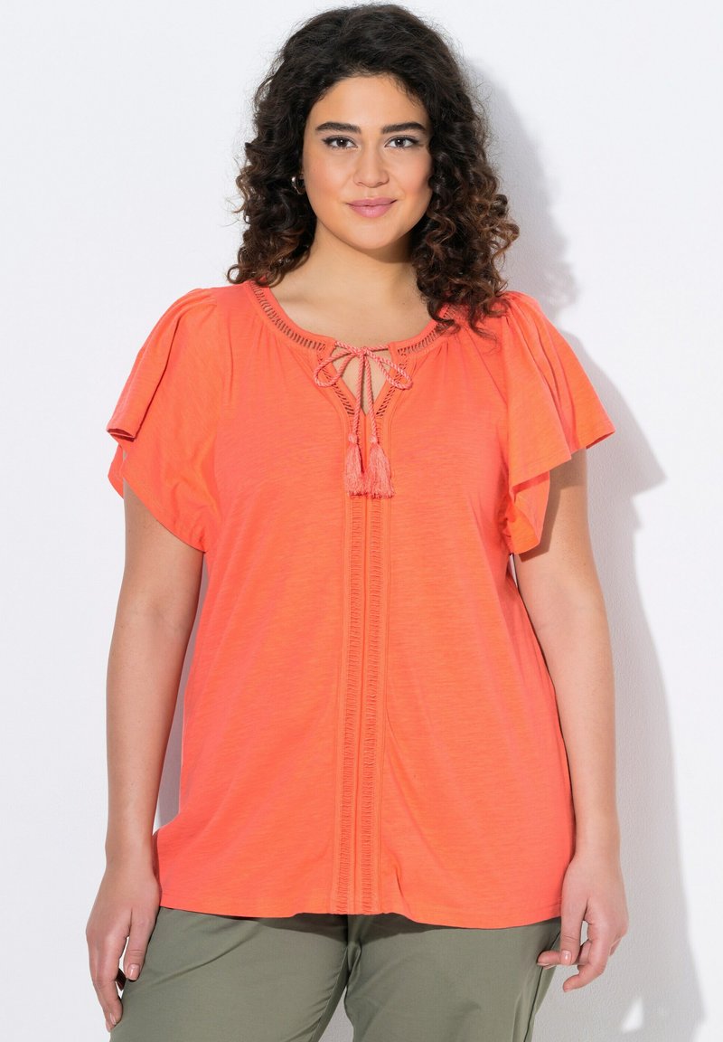 Korálová tunika s krátkým rukávem a kulatým výstřihem, zdobená třásněmi a vertikálním prošíváním. Měkká, lehká tkanina s volným střihem.