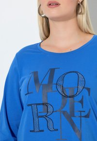 Niebieska koszula z długim rękawem, ozdobiona dużymi czarnymi literami i błyszczącym okrągłym akcentem. Miękka, teksturowana tkanina z okrągłym dekoltem.