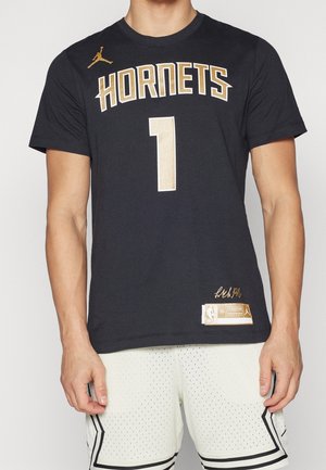 Mężczyzna w czarnej koszulce koszykarskiej Hornets z numerem 1 i białych perforowanych szortach, stojący ze zrelaksowanymi rękami opuszczonymi wzdłuż ciała.