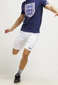 Marineblaues T-Shirt mit einem Wappenlogo, weiße Sportshorts und schwarze Turnschuhe. Graue Socken mit einem Musterakzent. Dynamische Pose.