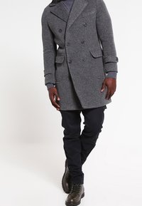 Abrigo de mezcla de lana gris con patrón texturizado, con solapas oversized y dos bolsillos delanteros, combinado con pantalones negros y botas marrones.