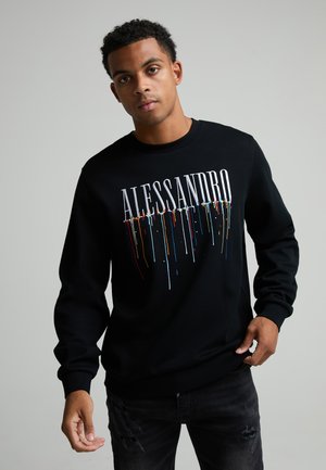Jeune homme portant un sweat-shirt noir avec le texte "ALESSANDRO" et des gouttes de peinture colorées, debout devant un fond uni gris clair.