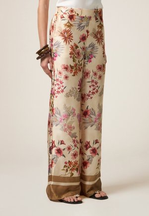 Beige, vidbenede satinbukser med flerfarvet blomsterprint og brun kant, båret med sorte sandaler og brune armbånd på venstre håndled.