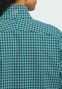 Teal og sort ternet bluse med en struktureret krave. Har et lille lilla logo på kraven. Vevet stofstruktur er synlig.